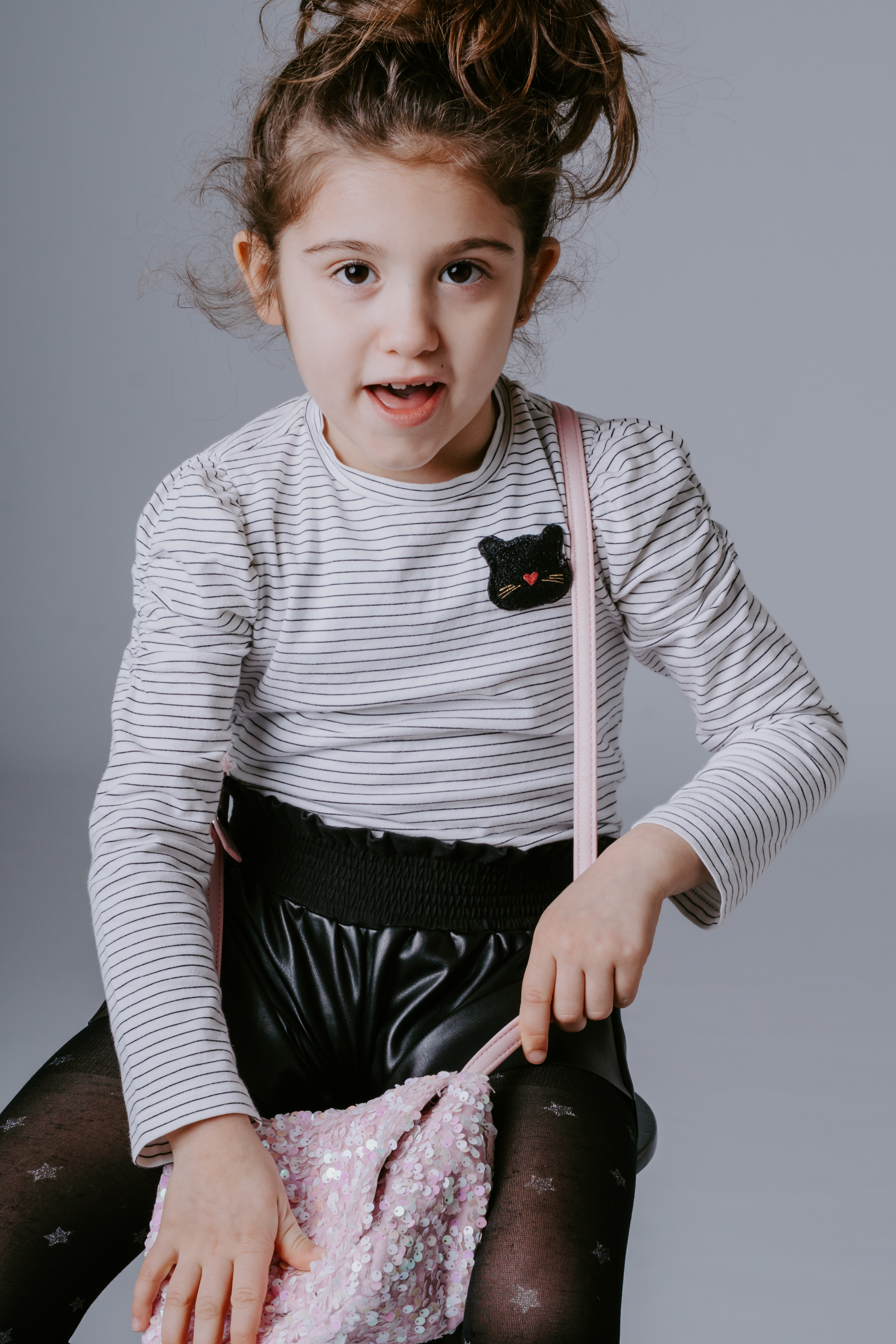 Bambina seduta su un cubo bianco che ride con gli occhi chiusi, con una borsetta rosa appoggiata accanto a lei.