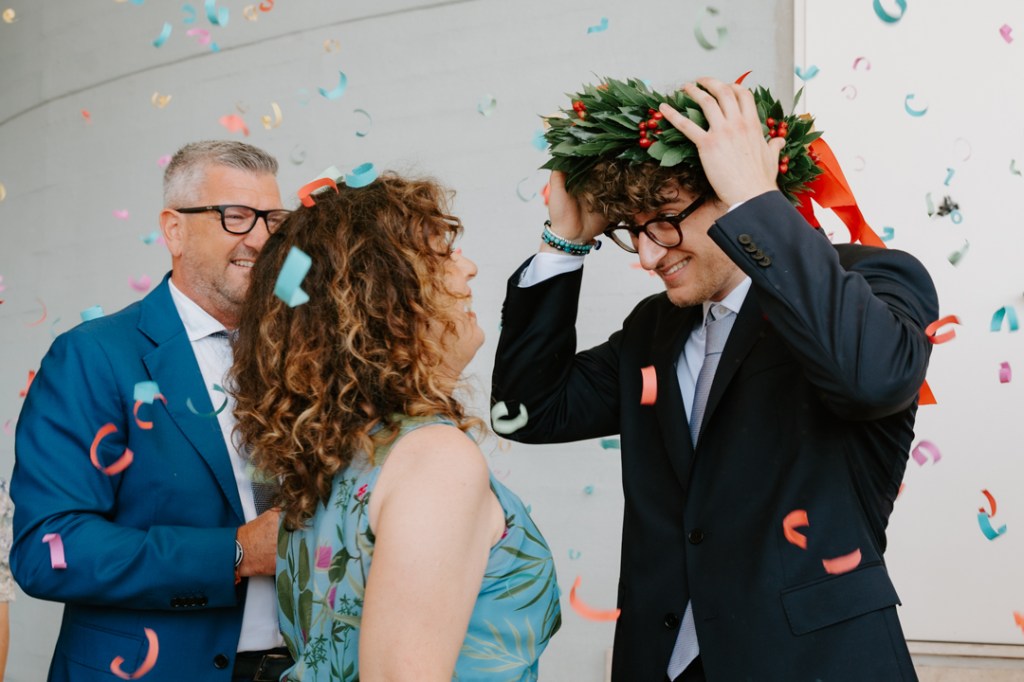 Giovane laureato indossa la corona d’alloro circondato dalla famiglia, tra sorrisi e coriandoli colorati, davanti all’ospedale di Cattinara a Trieste.